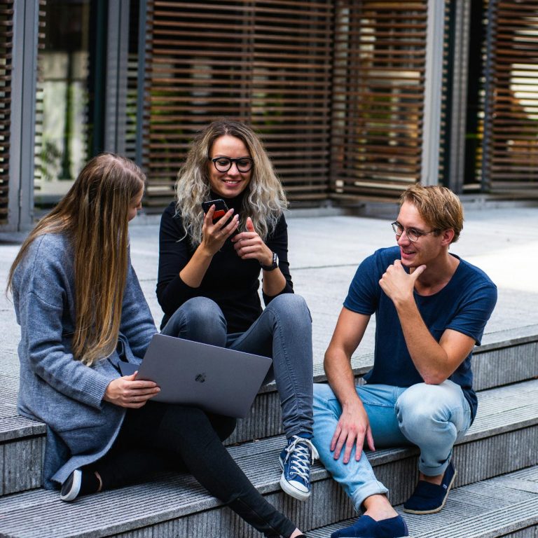 Zwei junge Frauen und ein Mann sitzen vor einem modernen Gebäude.